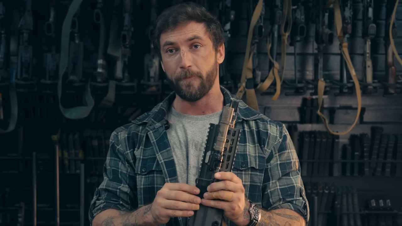 Man in plaid shirt holding a rifle part with a wall of firearms in the background