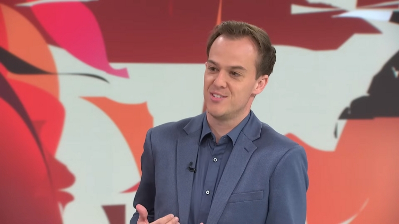 Guy Heveldt in a blue blazer speaking on a TV set with a red and white background