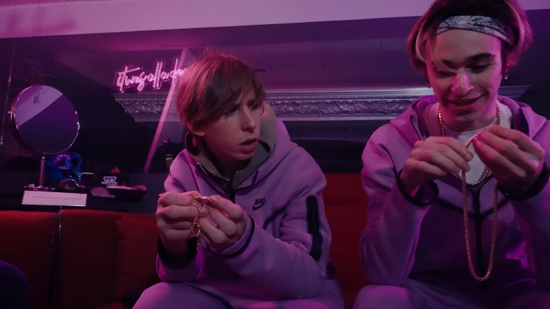 Two young men in matching purple tracksuits sit on a couch under neon lights, examining gold chains