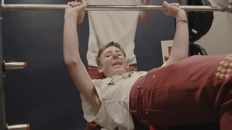 Lil Maceee lies on a bench press, lifting a barbell with a spotter standing behind him