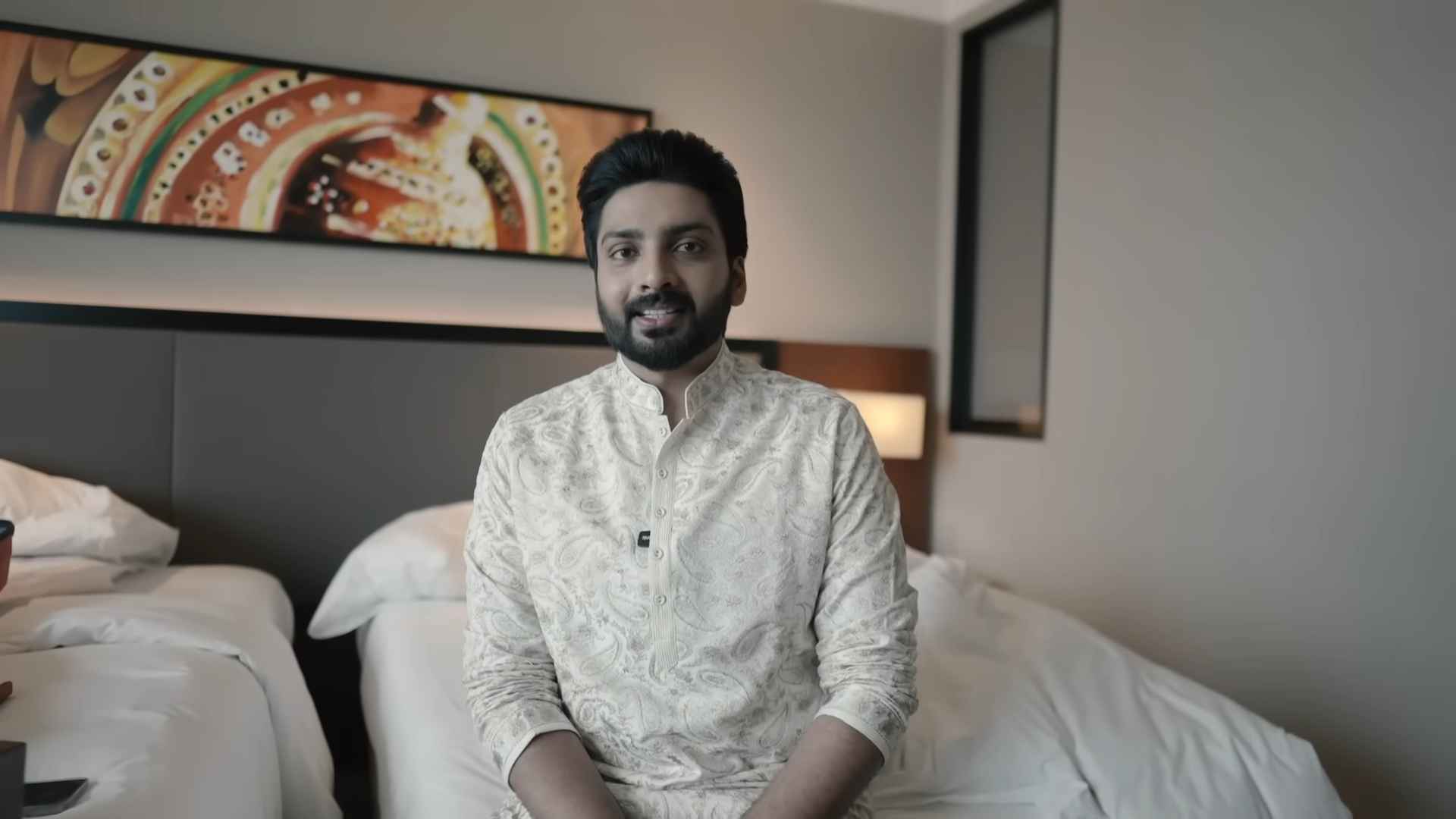 Thejus Jyothi sitting on a bed in a hotel room, wearing a cream-colored traditional kurta