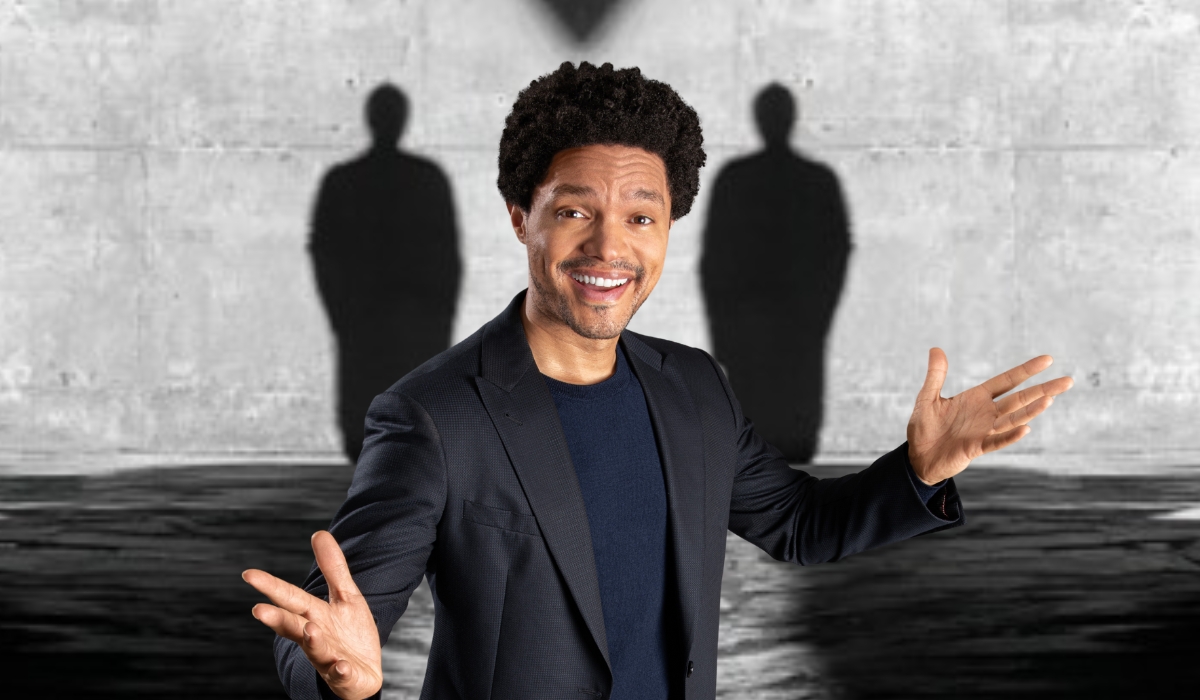 Man wearing a dark blazer and navy shirt smiles with open arms in front of a gray wall showing two human-shaped shadows