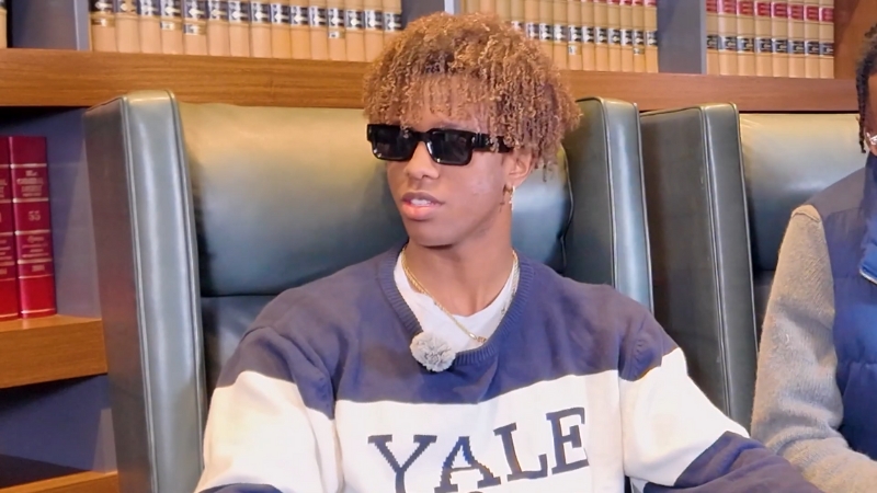Nathaneo Johnson wearing a Yale sweatshirt and sunglasses sits in a library setting during an interview