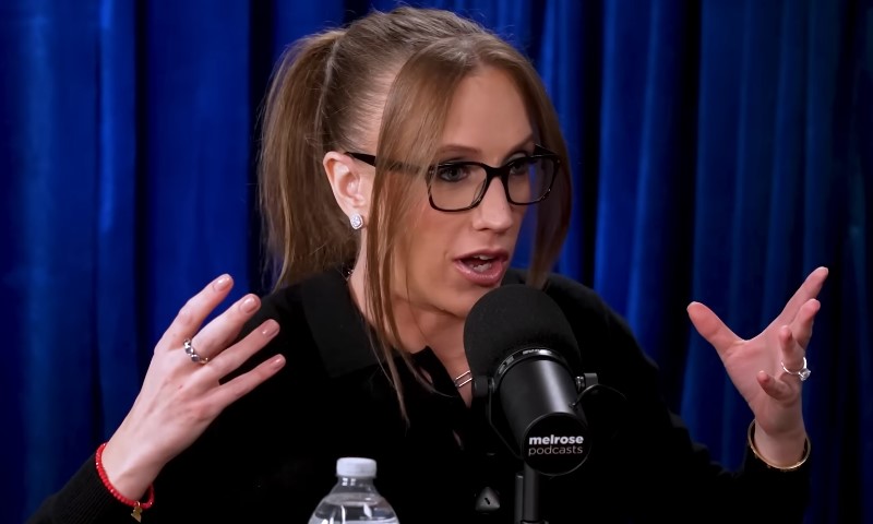 A woman with glasses, Kat Timpf, is speaking into a microphone during a broadcast