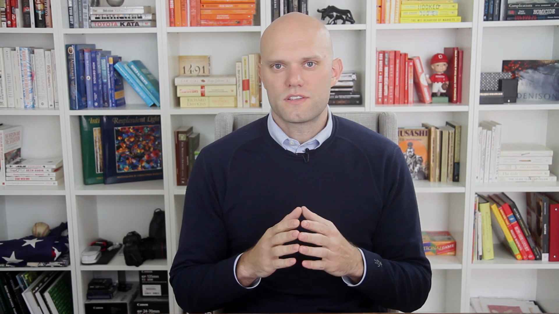 A person sitting in front of a bookshelf, speaking or explaining something with hands together