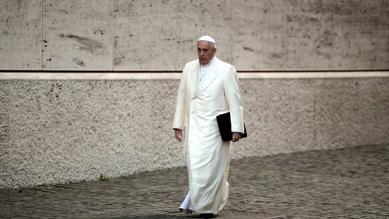 Pope Francis walking in white papal robes carrying a folder