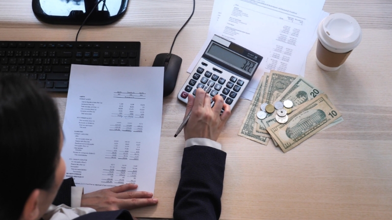 A person reviews printed financial reports with cash on the desk and types on a calculator