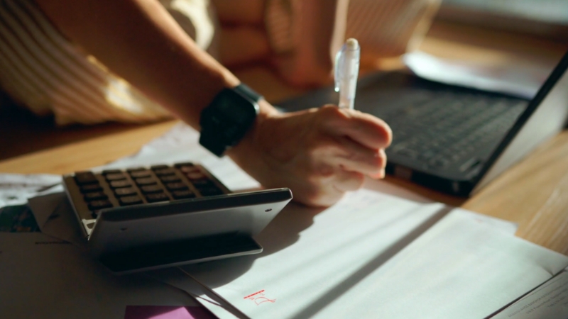 A person works with a calculator and paperwork spread out on a desk to review the real costs needed to keep a business running