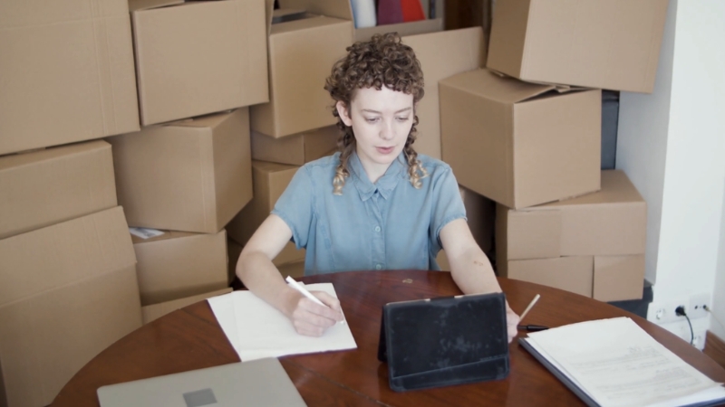 A small business owner works at a table surrounded by stacked shipping boxes