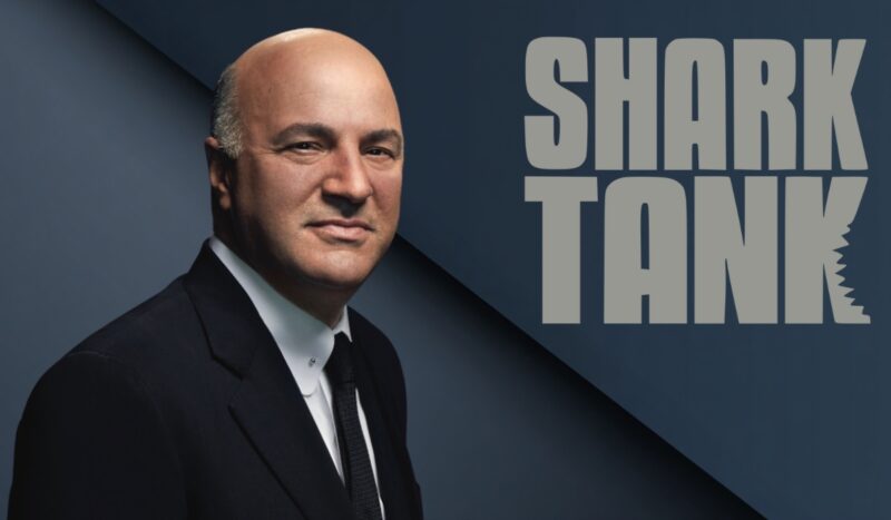 A Shark Tank investor in a suit posing beside the show logo