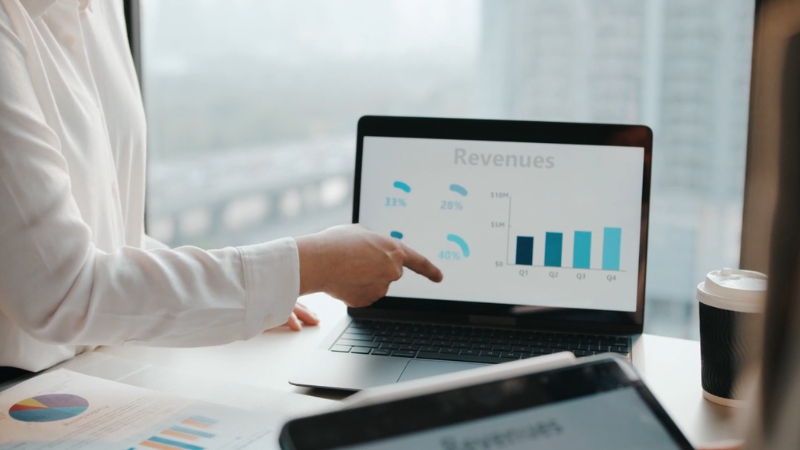 Person pointing at revenue charts on a laptop screen during a desk meeting