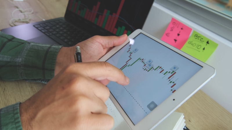 Hands using a tablet that displays financial charts and market data on a desk with notes and a laptop in the background