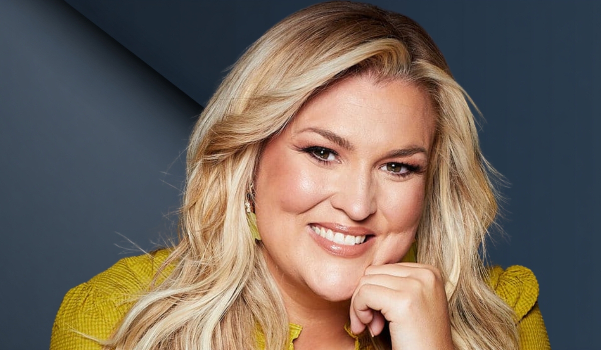 A professional headshot of Sara Davies smiling with her hand resting under her chin, wearing a yellow textured top against a blue-gray background