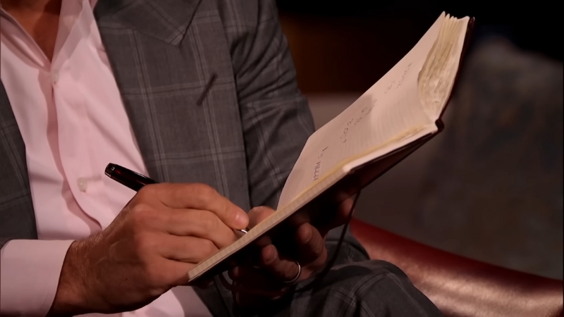 Investor in a suit taking notes in a notebook during a Shark Tank pitch