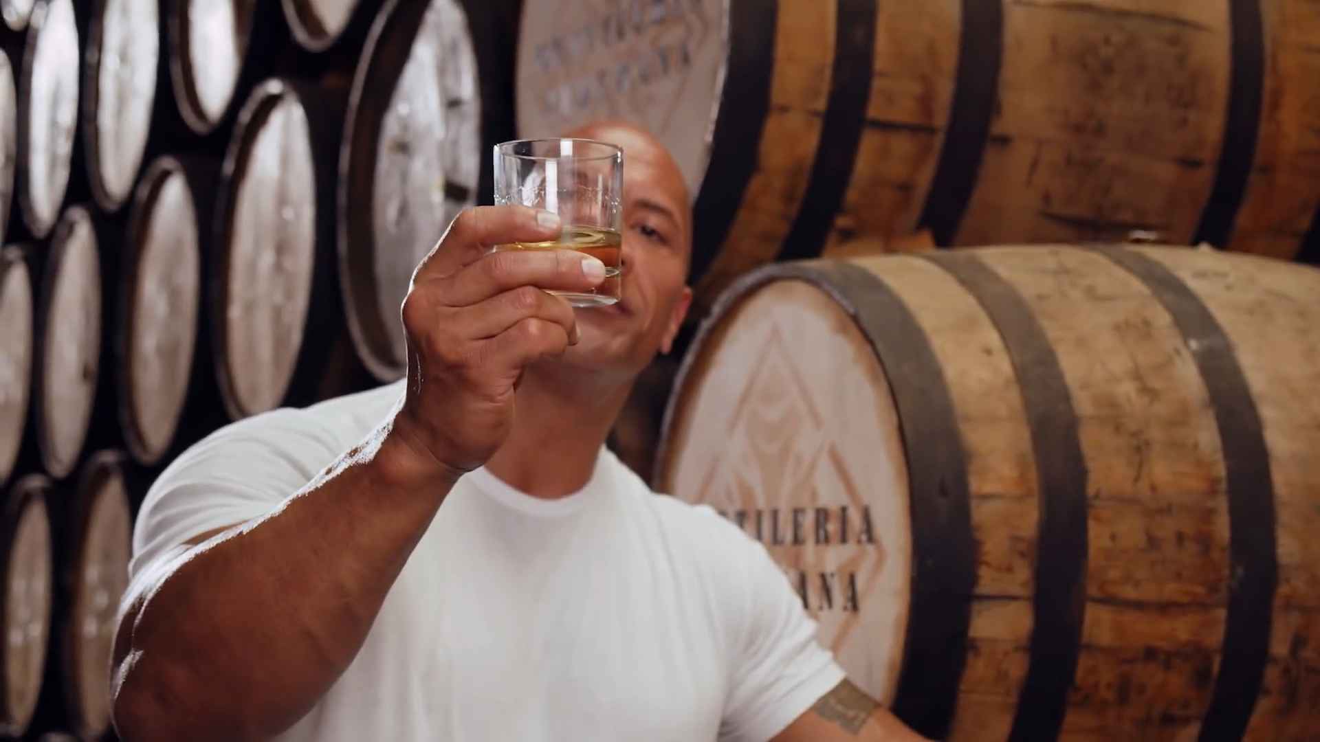 Man holding a glass of tequila in front of wooden barrels inside a distillery