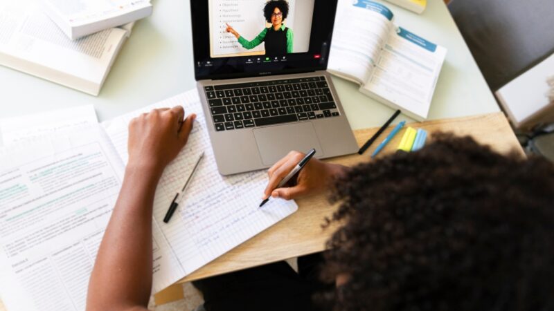 Student follows an online class on a laptop while taking notes at a desk