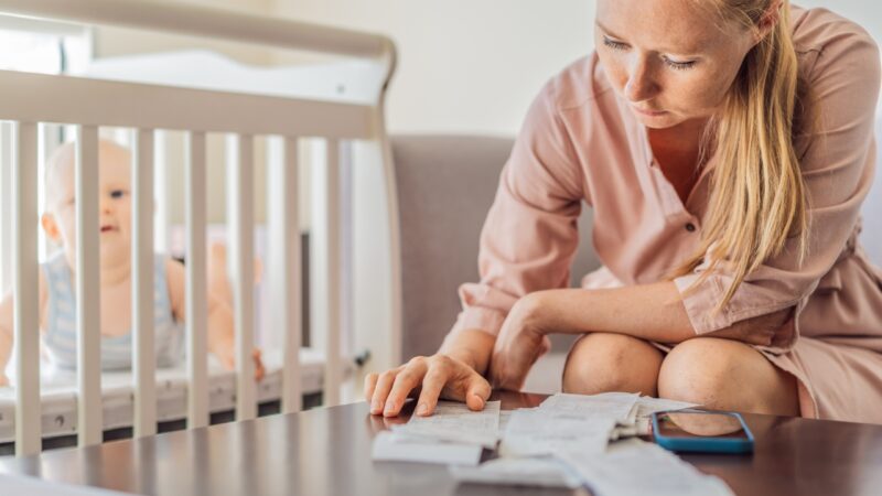 A mother reviews childcare bills while her baby sits safely in a crib nearby